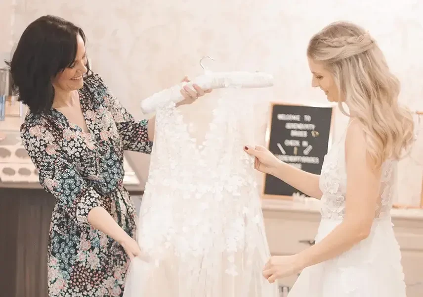 Two women smile while holding a white wedding dress on a hanger in a warmly lit bridal shop. A sign in the background adds a welcoming touch.