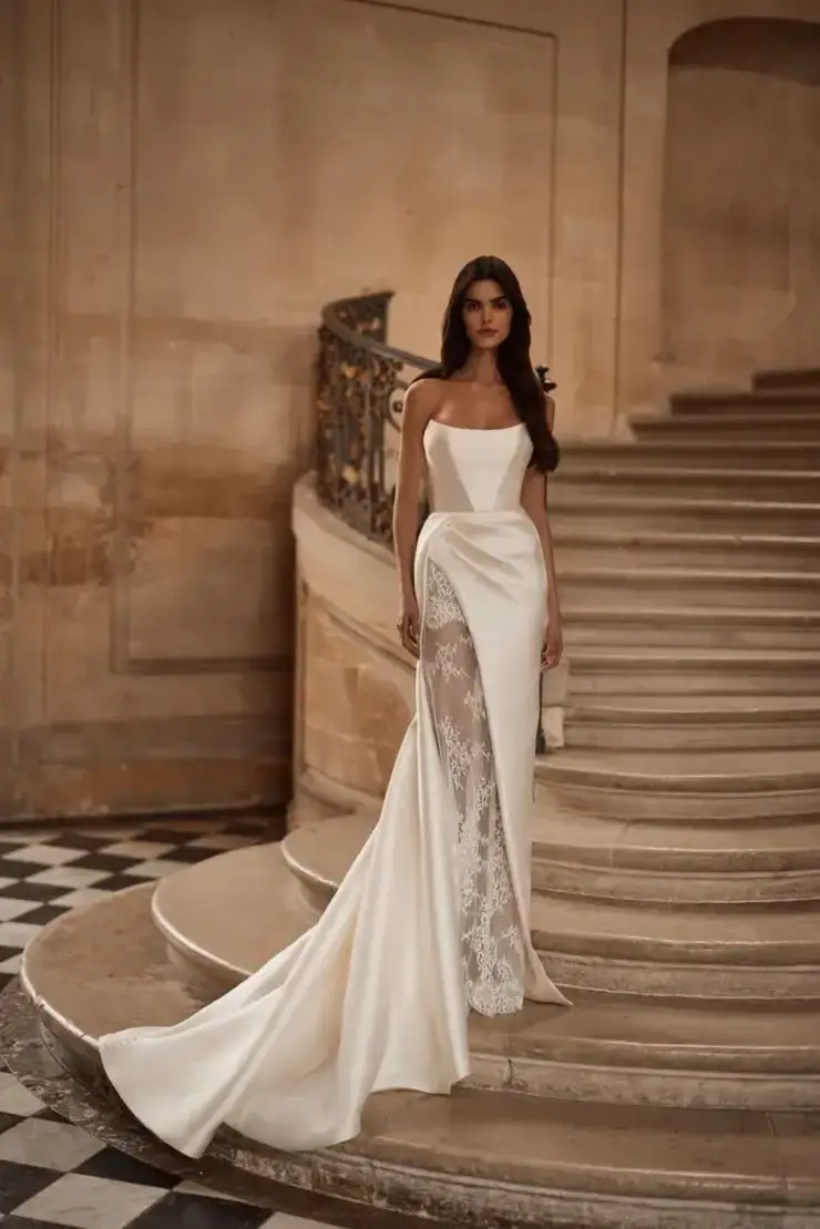 A woman in an elegant white gown with lace details stands on a grand stone staircase in a stately setting, conveying sophistication and grace.