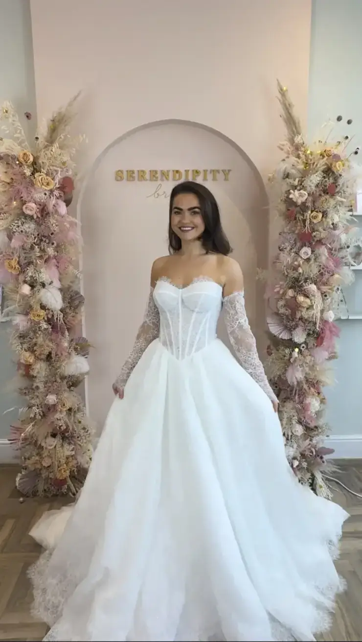 A bride in a white lace wedding dress smiles in front of a floral archway, with pastel flowers and pampas grass, under a 