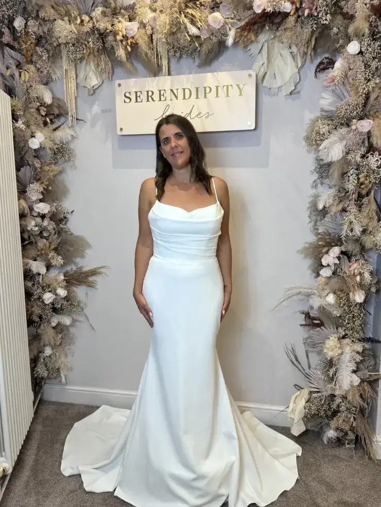 A woman in a white wedding dress stands in front of a decorated backdrop featuring floral arrangements and a sign that reads 