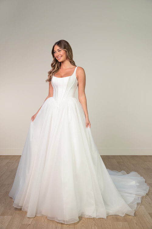 A smiling woman in a flowing, sleeveless, white wedding dress with a fitted bodice stands on a wooden floor against a plain background, exuding elegance.