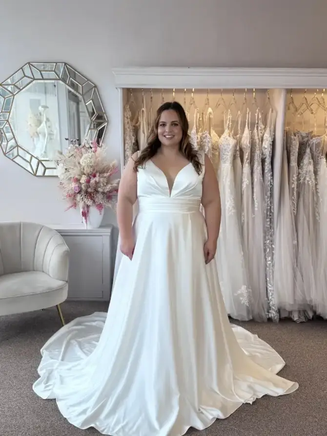 A woman wearing a white wedding dress stands in a bridal shop, surrounded by wedding dresses displayed on racks. There is a decorative mirror and a floral arrangement in the background.