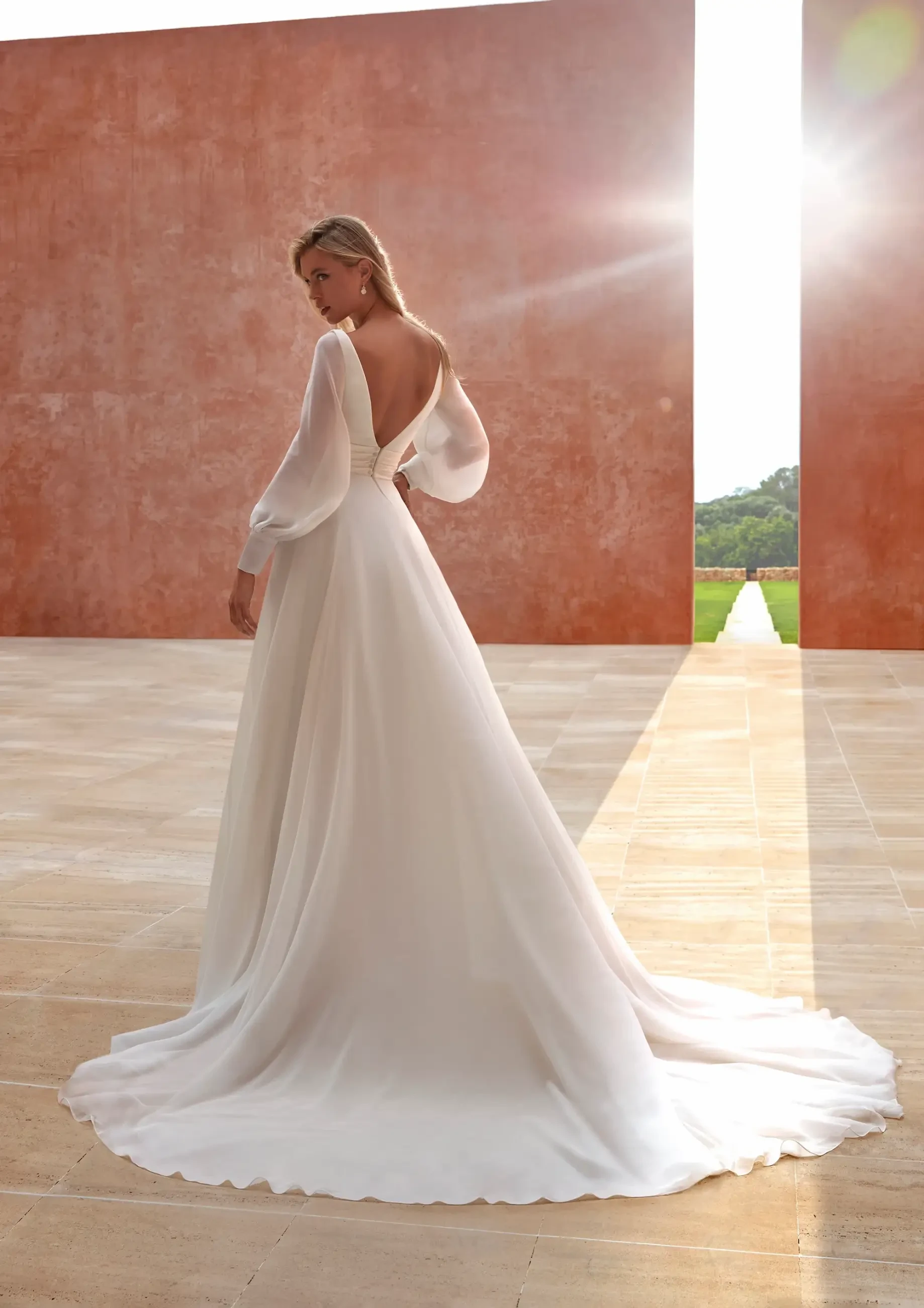 A woman in an elegant white wedding gown with sheer long sleeves stands outdoors. Sunlight streams between two terracotta walls, casting a warm glow.