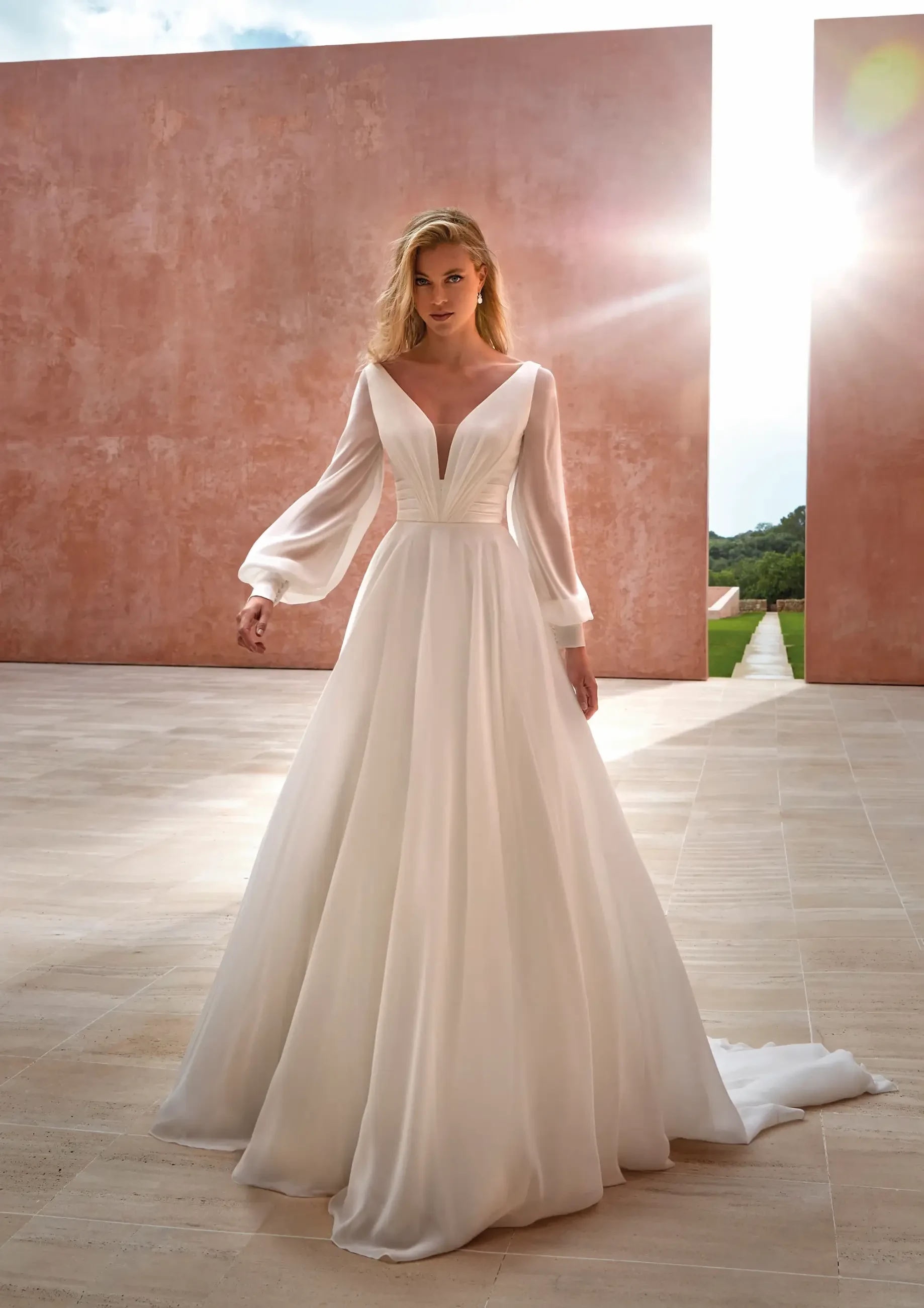 A woman in an elegant white wedding gown with sheer long sleeves stands outdoors. Sunlight streams between two terracotta walls, casting a warm glow.