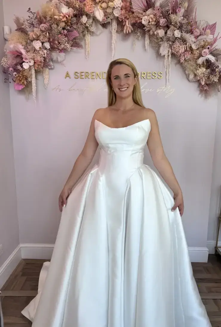 A woman wearing a strapless white wedding gown stands in front of a floral backdrop.
