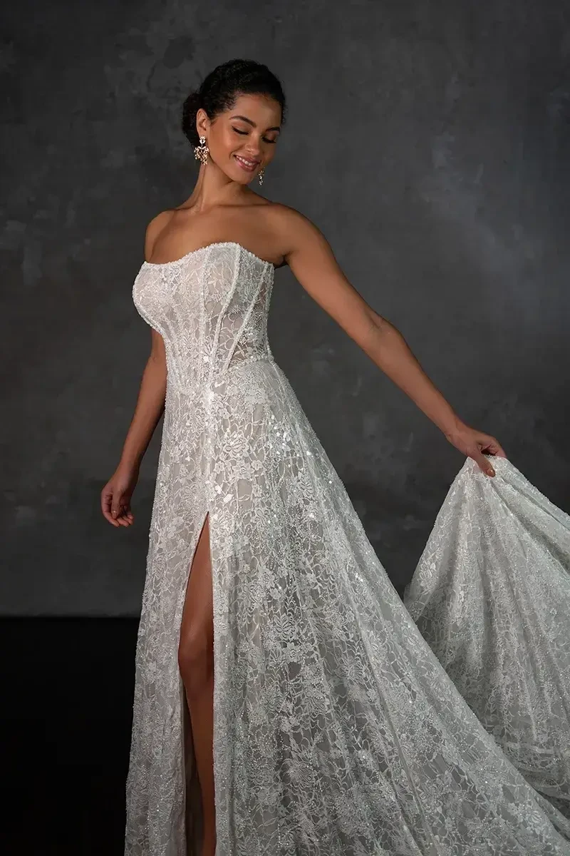 Model is wearing a strapless sheath gown with sparkle lace, a soft scoop neckline, corset-style bodice, and high leg slit.
