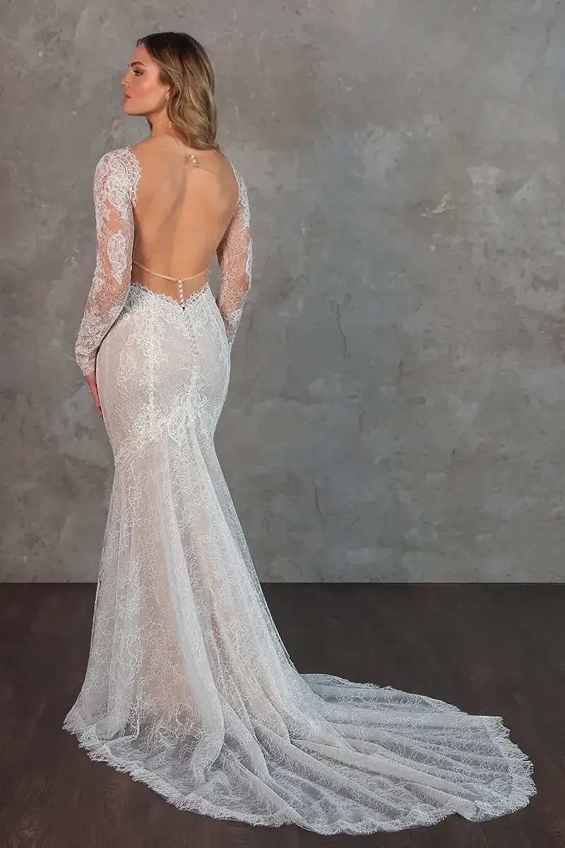 A woman in a lace wedding gown with long sleeves and a plunging neckline stands against a textured gray wall. She looks poised and elegant.
