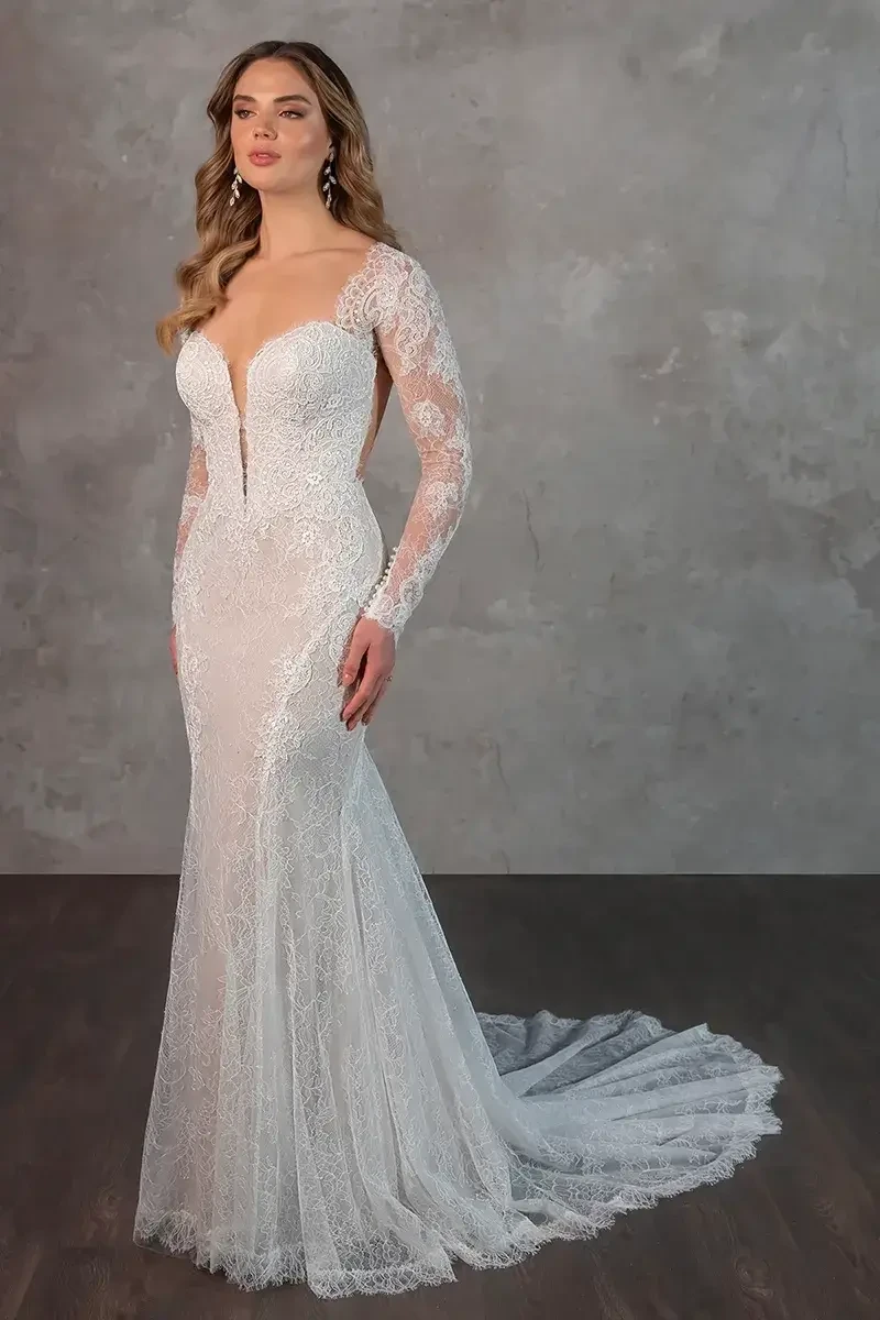 A woman in a lace wedding gown with long sleeves and a plunging neckline stands against a textured gray wall. She looks poised and elegant.