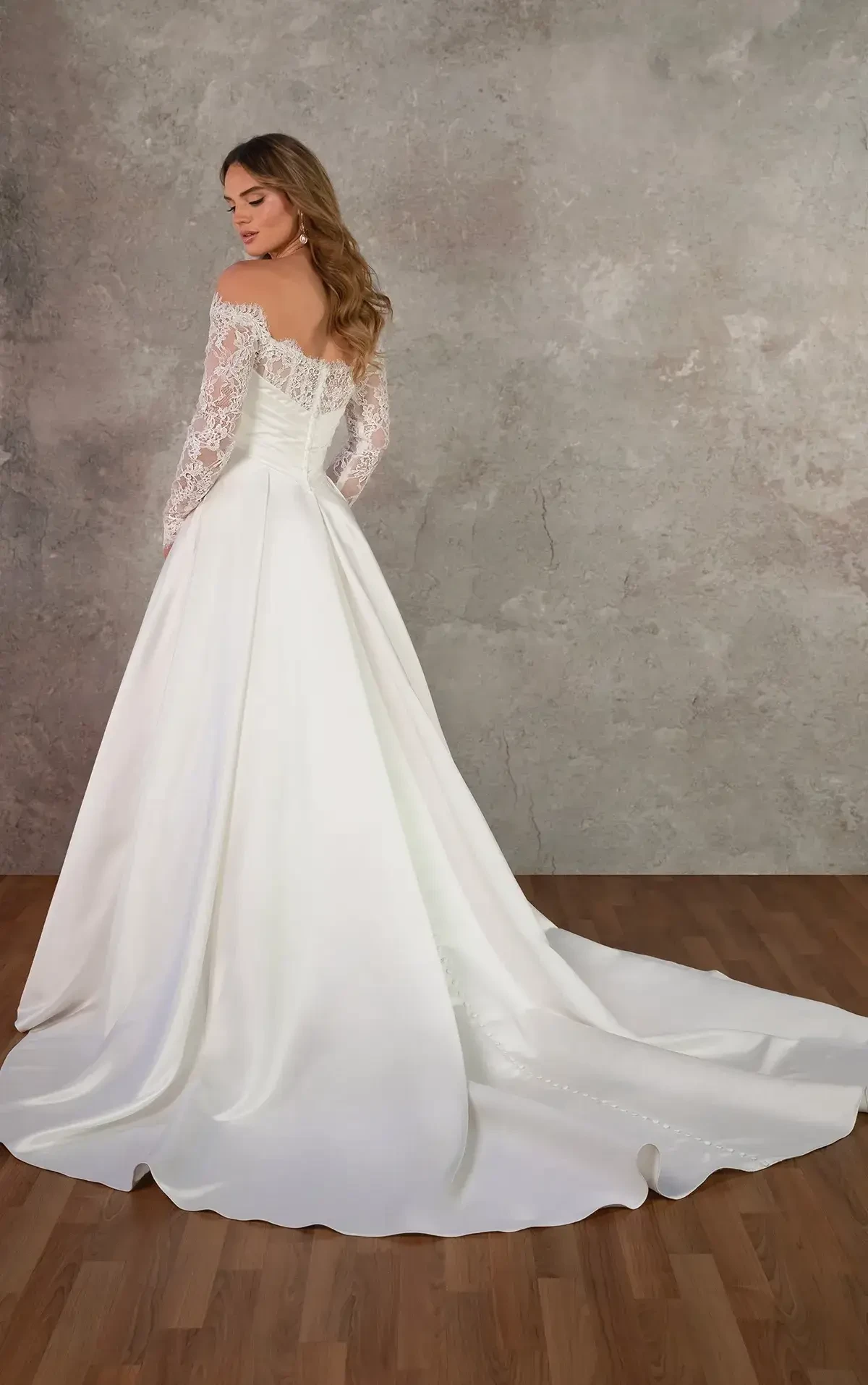 A woman in a flowing white dress with lace sleeves gracefully descends a wooden staircase. Behind her is a mural of a tranquil pastoral scene.
