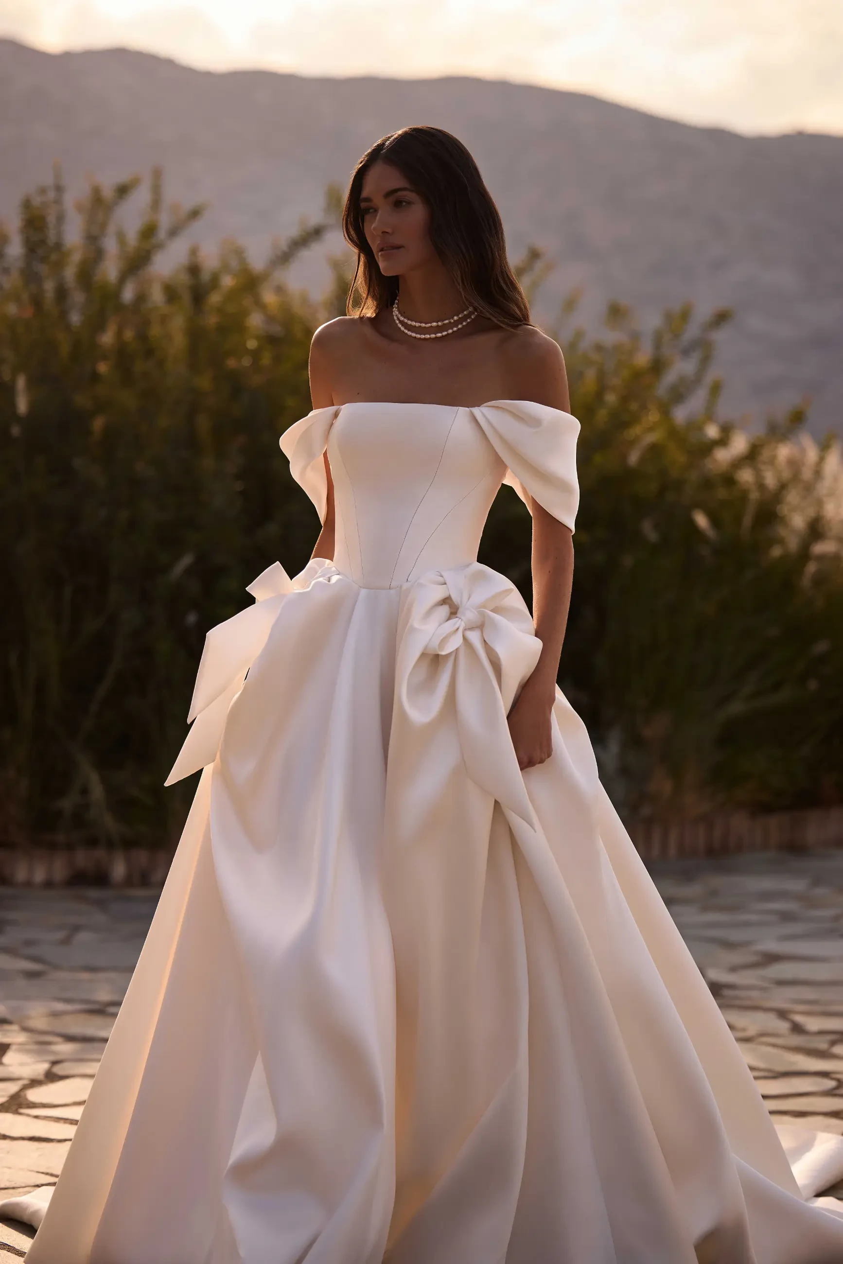 Bride in elegant off-shoulder ivory gown with large bow, standing outdoors at sunset. Mountain backdrop, warm tones, serene ambiance.