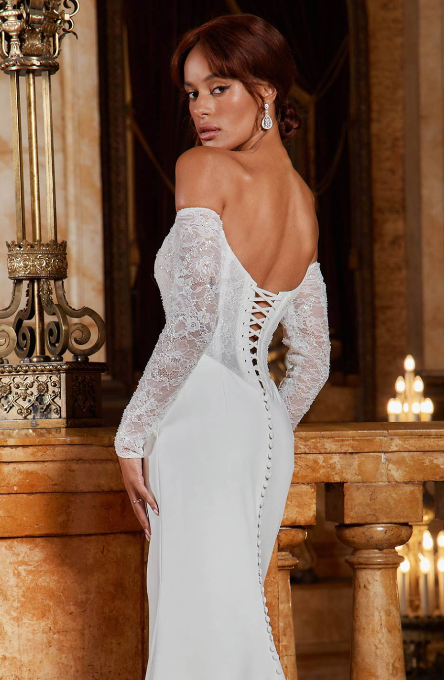 A woman in an elegant off-shoulder white gown with lace details stands in an opulent room with marble and candle-lit ambiance, conveying luxury and grace.