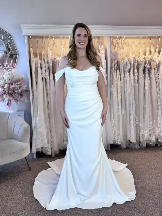 A woman in an elegant off-shoulder white wedding dress stands in a bridal shop. She smiles warmly, surrounded by more bridal gowns and a soft chair.