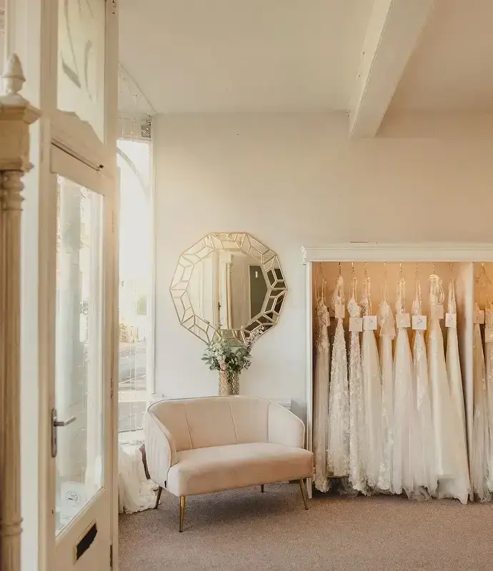 A bright and airy bridal shop interior featuring a plush pink sofa, a decorative round mirror on the wall, and a display of wedding dresses hanging in a row.