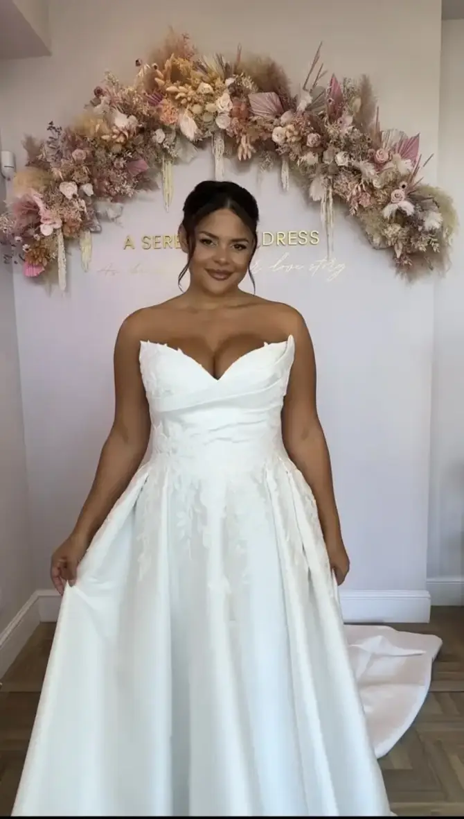 A woman wearing a white strapless wedding dress stands in front of a floral backdrop.