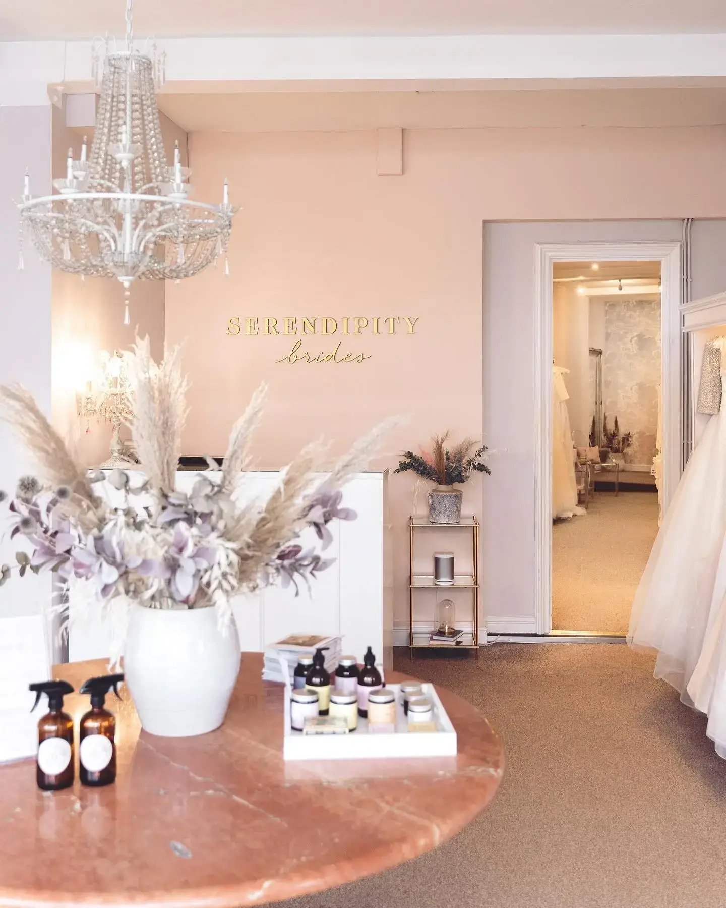 Interior of a boutique with soft pink walls, decorative plants in a white vase on a round wooden table, and a chandelier hanging from the ceiling. A sign reads 